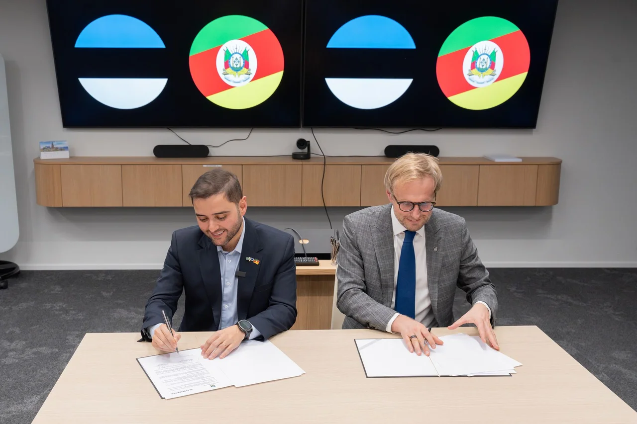 Two men signing documents in front of a country flag wall.